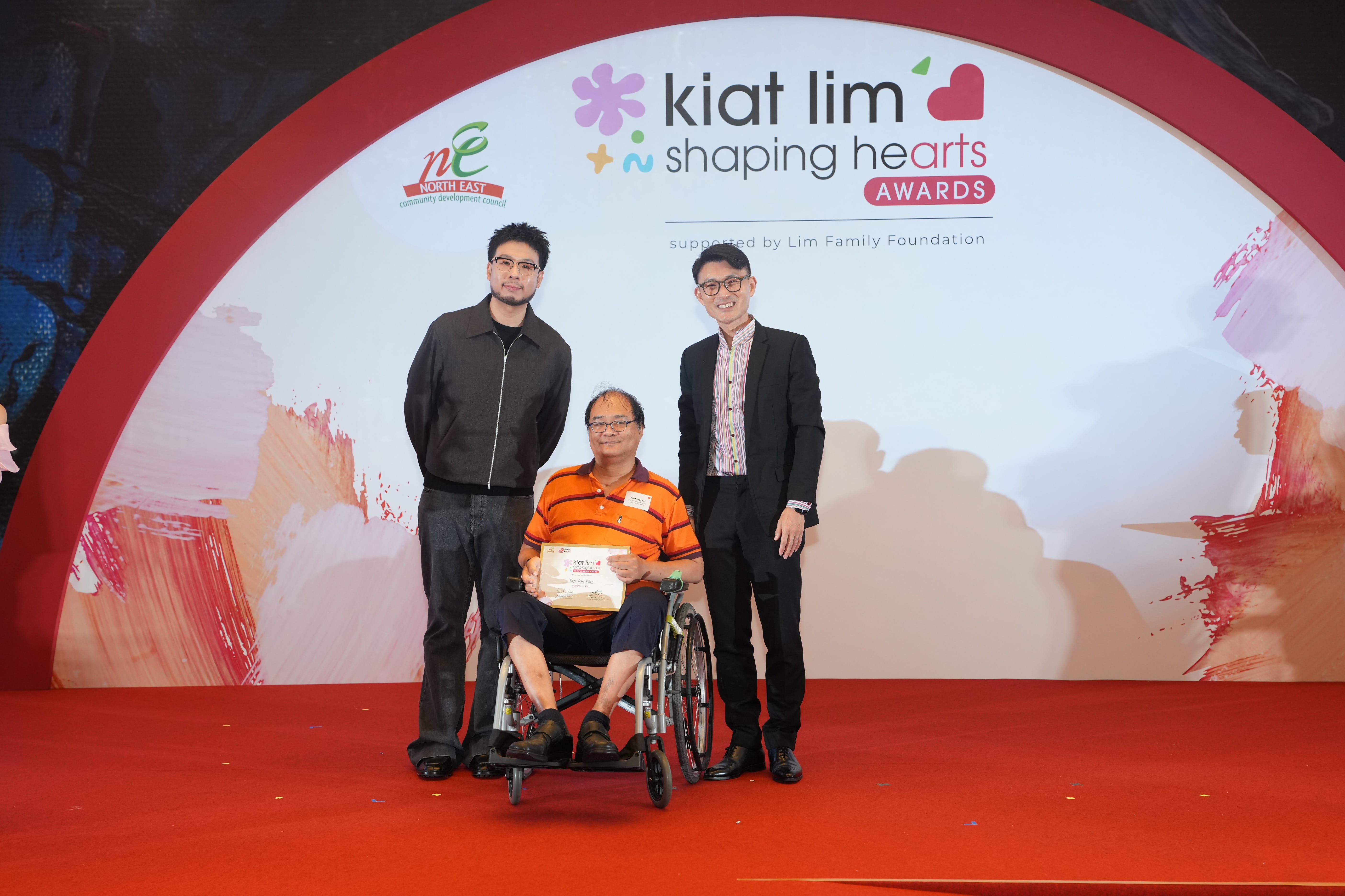 Award recipient standing on stage with the presenter, holding their certificate and posing for a group photo during the award ceremony, with a backdrop reading ‘Kiat Lim-Shaping Hearts Awards'.